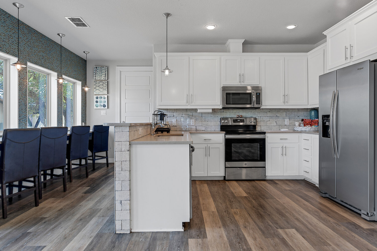 Modern white kitchen with stainless steel appliances, light gray subway tile backsplash, a small white peninsula, and an adjacent dining area with a bold blue and teal mosaic accent wall.