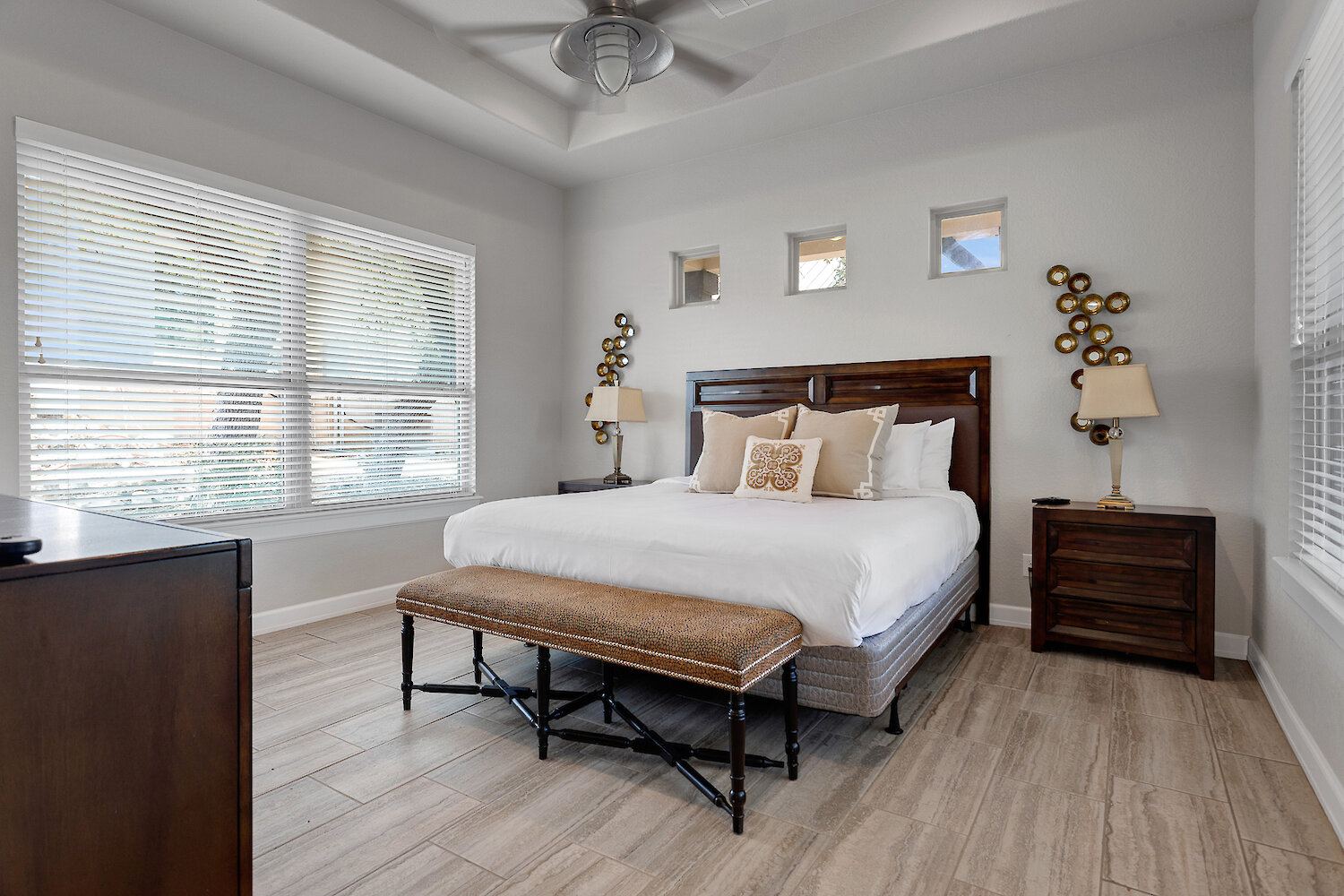 Modern bedroom with a dark wood king-size bed, upholstered animal print bench, contemporary metallic wall art, and light wood-look tile flooring.