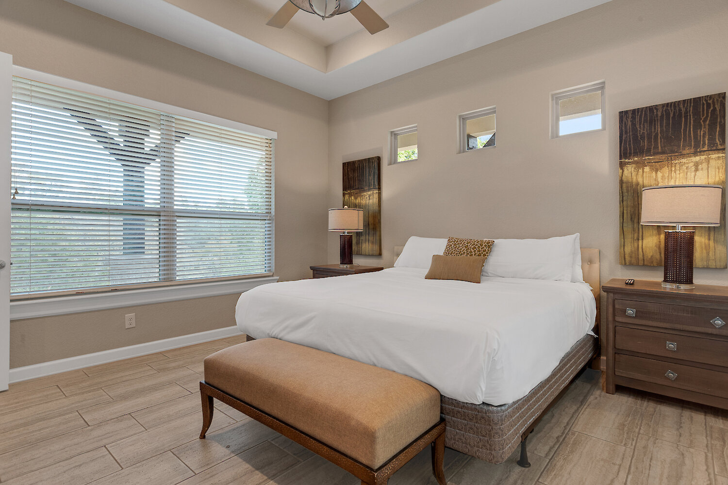 Cozy bedroom with a king-size bed, white linens, tray ceiling, dark wood nightstands with lamps, and large modern abstract art panels on both sides of the bed.