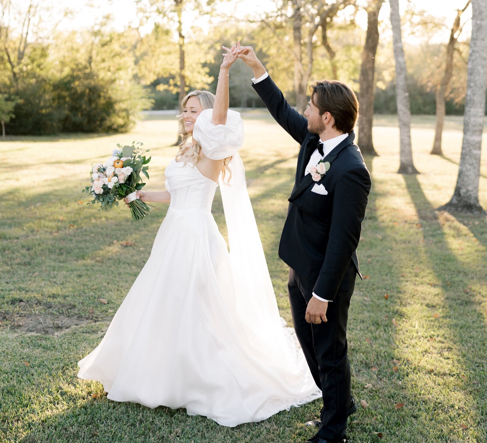 Bright outdoor portrait of a joyful bride and groom dancing in a park or field at sunset, with the bride holding a bouquet and wearing a dress with puff sleeves