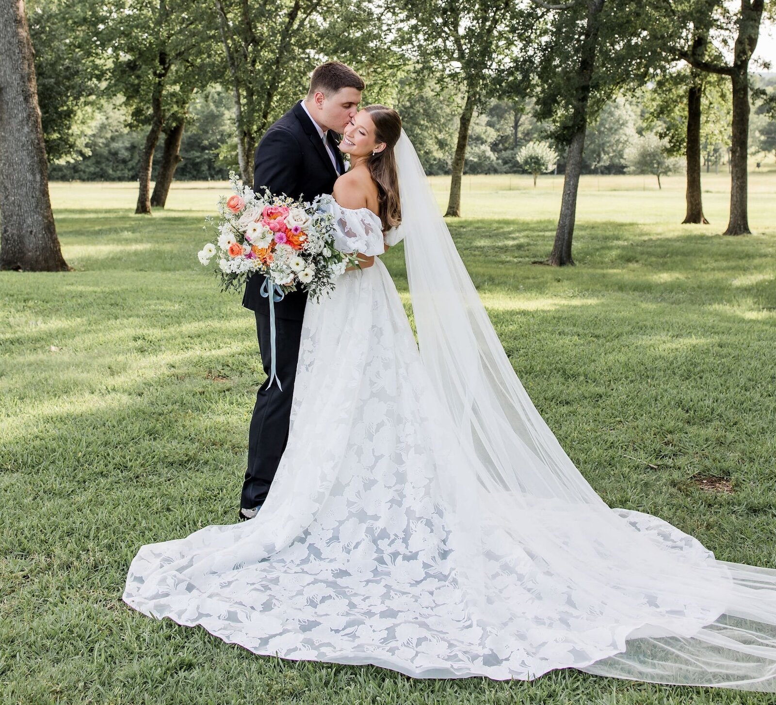 Joyful bride and groom embracing in a park or field, with the groom kissing the bride's cheek. The bride wears an off-the-shoulder lace dress with a long, sweeping veil and holds a colorful bouquet