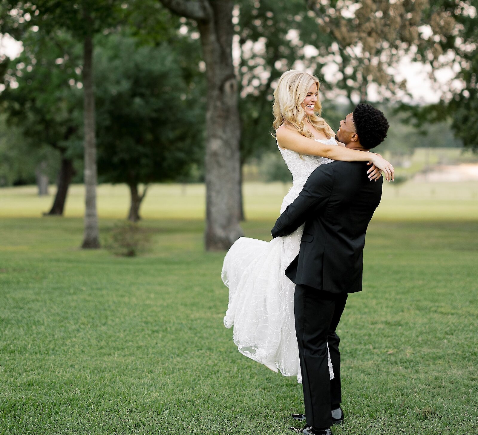 Joyful groom in a black suit lifting the bride in a white lace dress while embracing in a grassy park setting surrounded by tall trees at sunset.