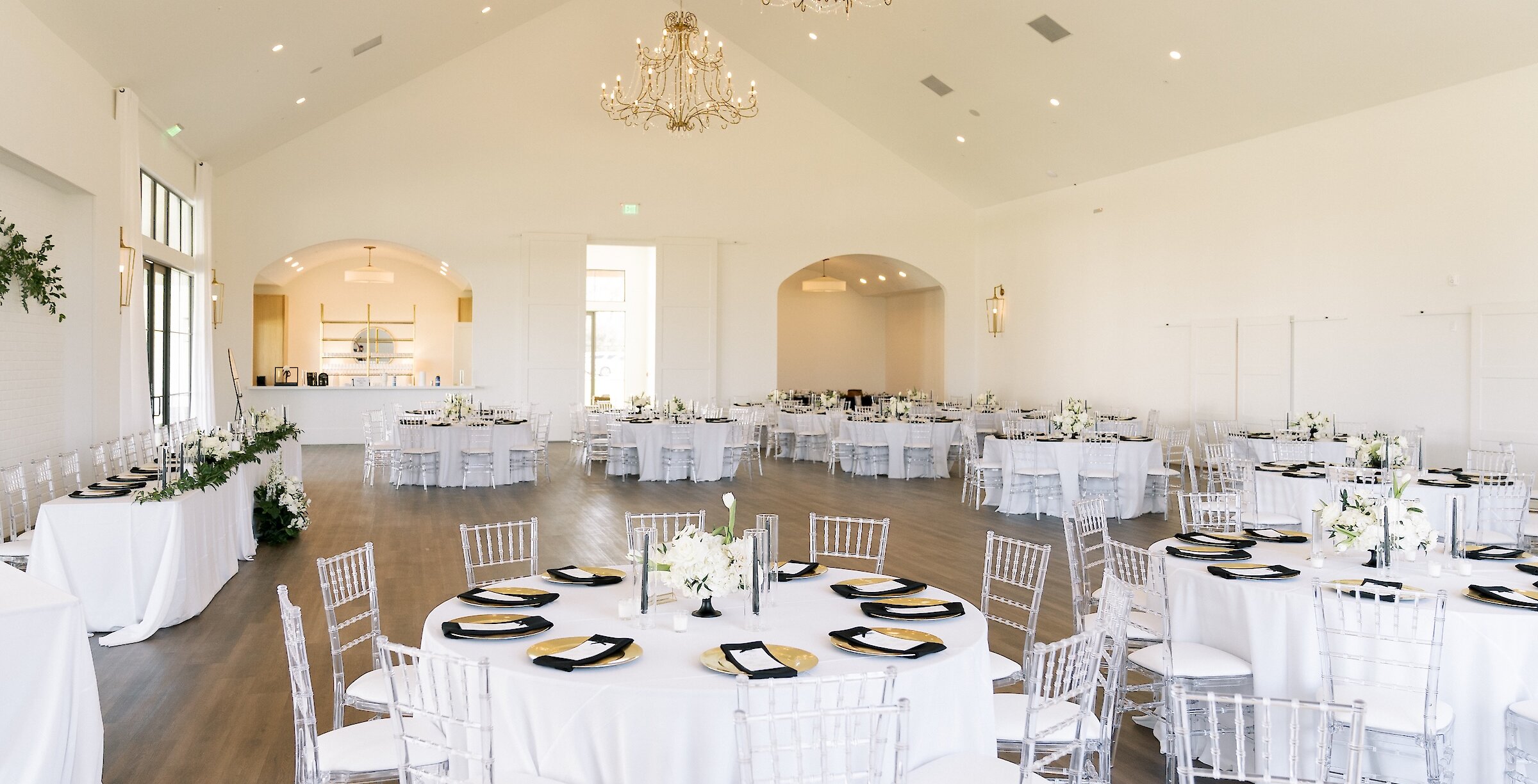 Spacious and elegant white wedding reception hall with vaulted ceilings, large crystal chandeliers, and white round tables set with clear acrylic chairs and gold chargers.