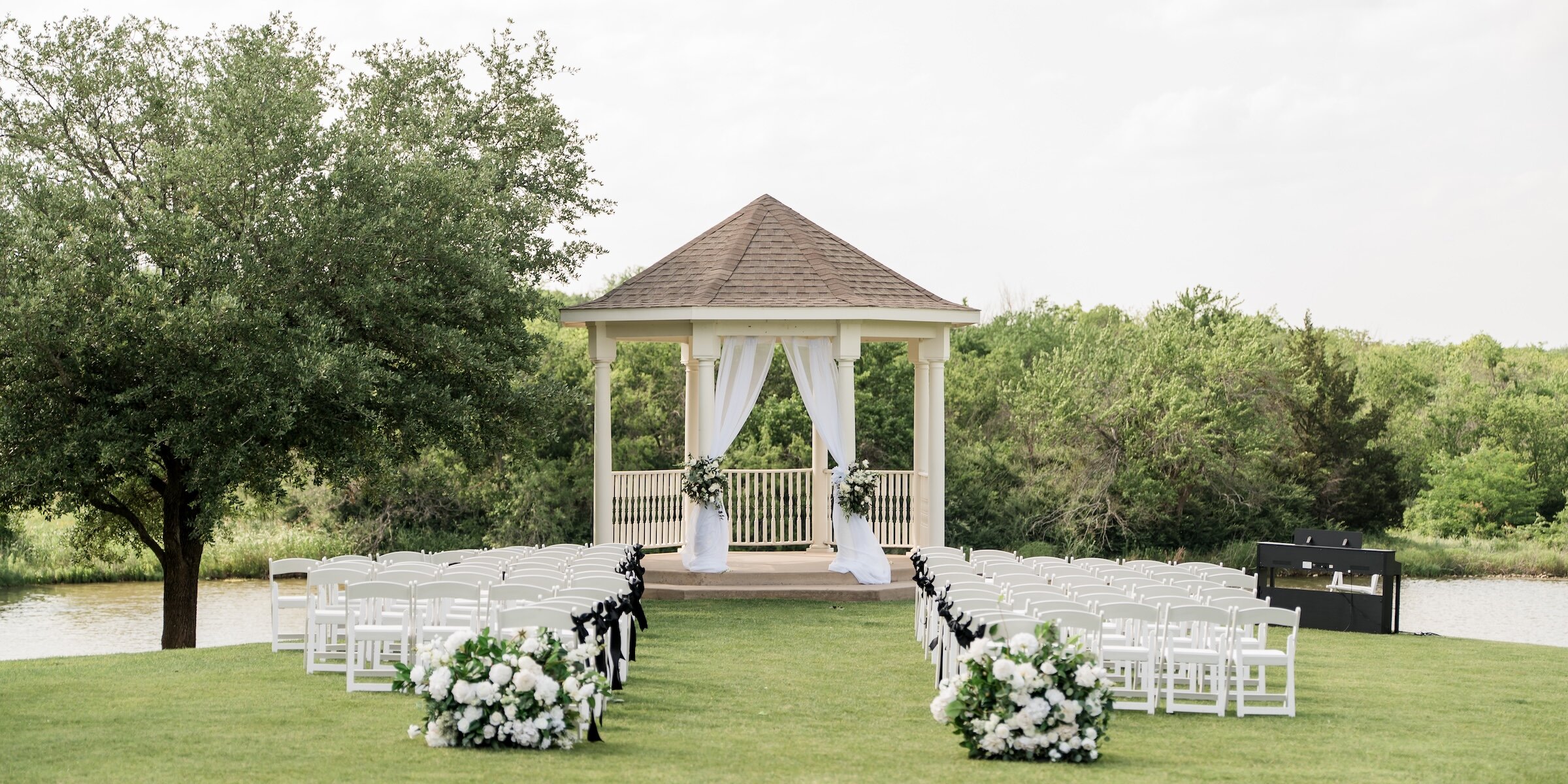 An outdoor wedding ceremony setup featuring rows of white chairs facing a white gazebo draped with sheer white fabric and flowers.