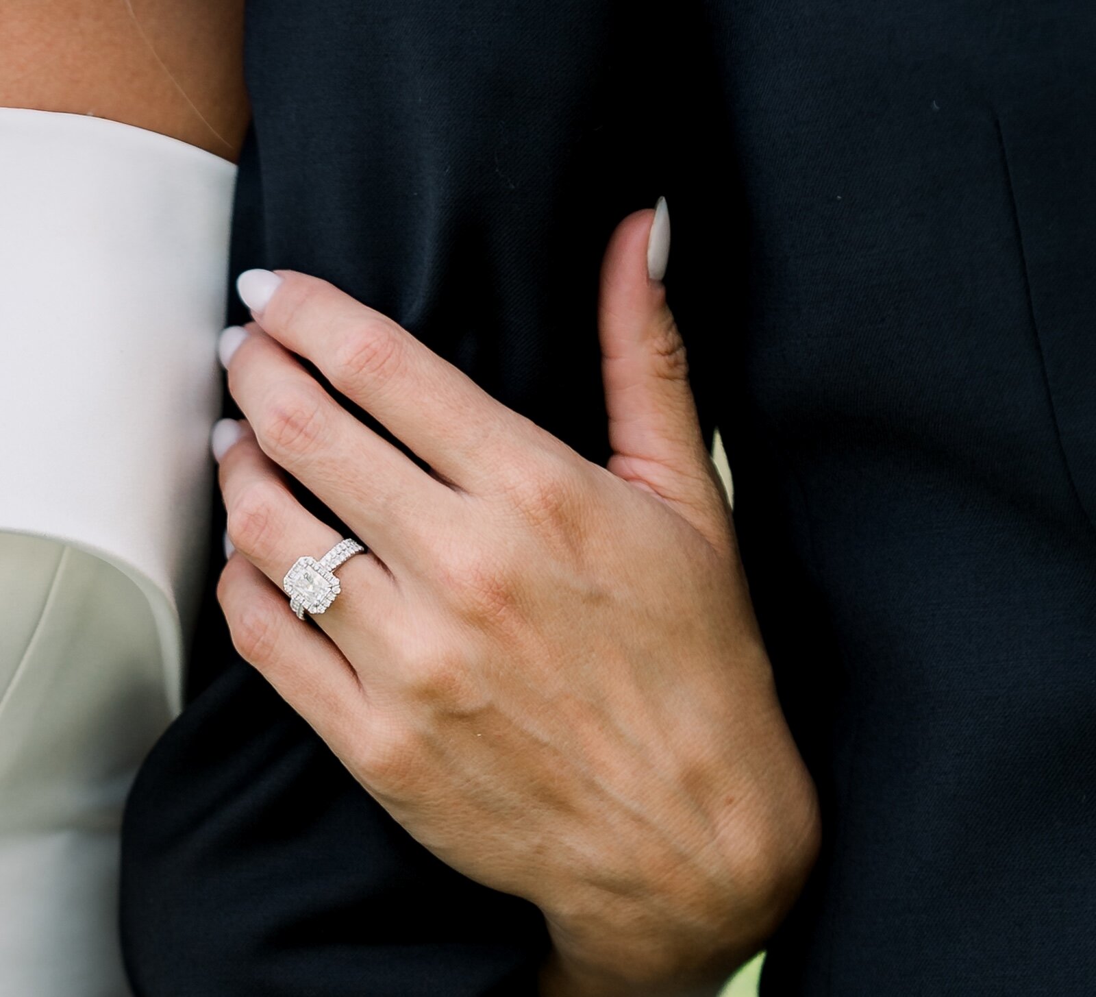 A close-up of a bride's hand with white nails resting on a groom's navy suit, showing a cushion-cut diamond engagement ring.
