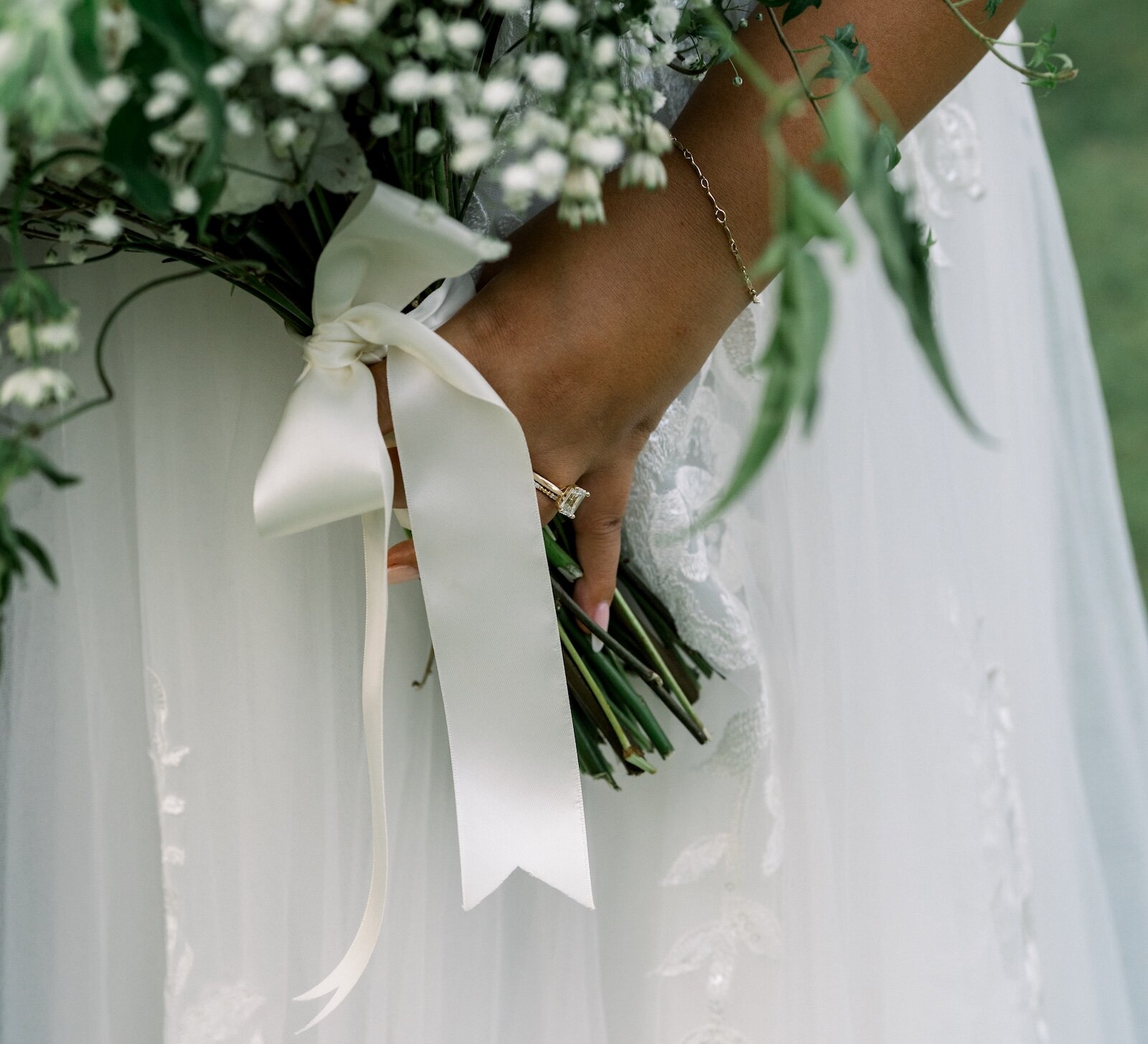 Close-up of a bride's hand holding a bouquet tied with a white ribbon, featuring a gold thin band engagement ring.