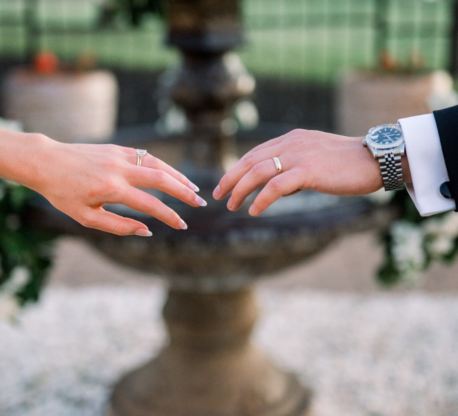 The hands of the bride wearing an engagement ring and groom reaching out over a decorative stone water fountain.