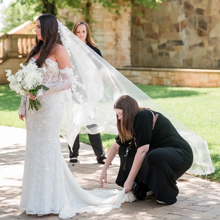 A bride in a lace wedding gown holding a white bouquet while two assistants adjust her long veil on a paved outdoor walkway.