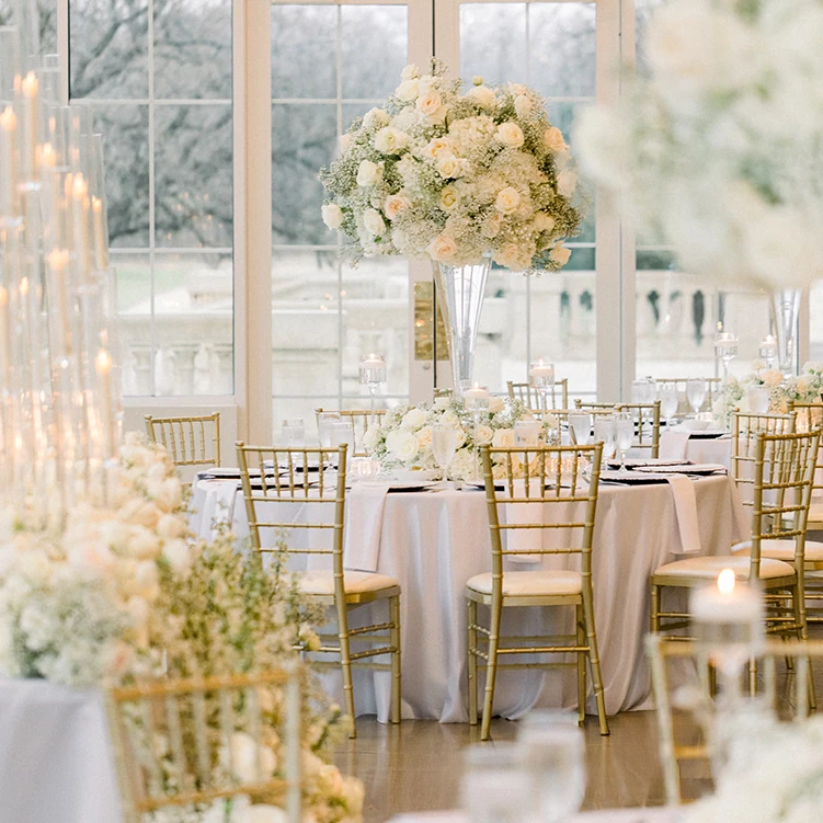 An elegant wedding reception table featuring gold chiavari chairs, white linens, and tall white floral centerpieces near large windows.