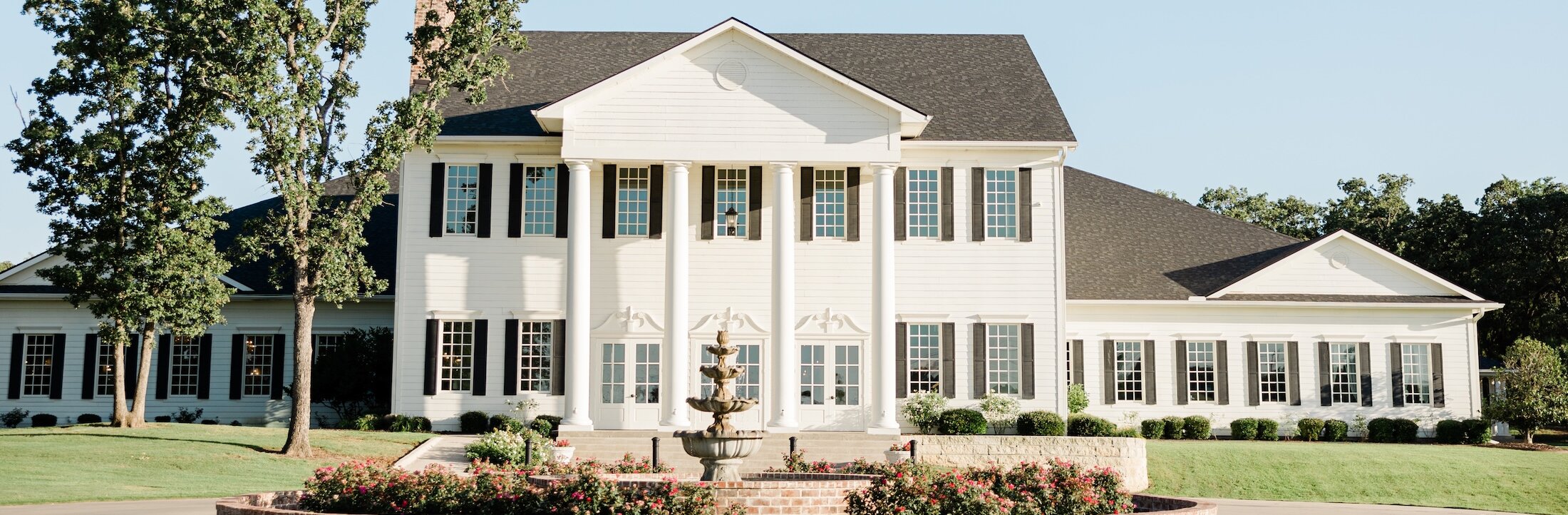 The exterior of a large, white, two-story Southern-style mansion with black shutters and a small fountain.