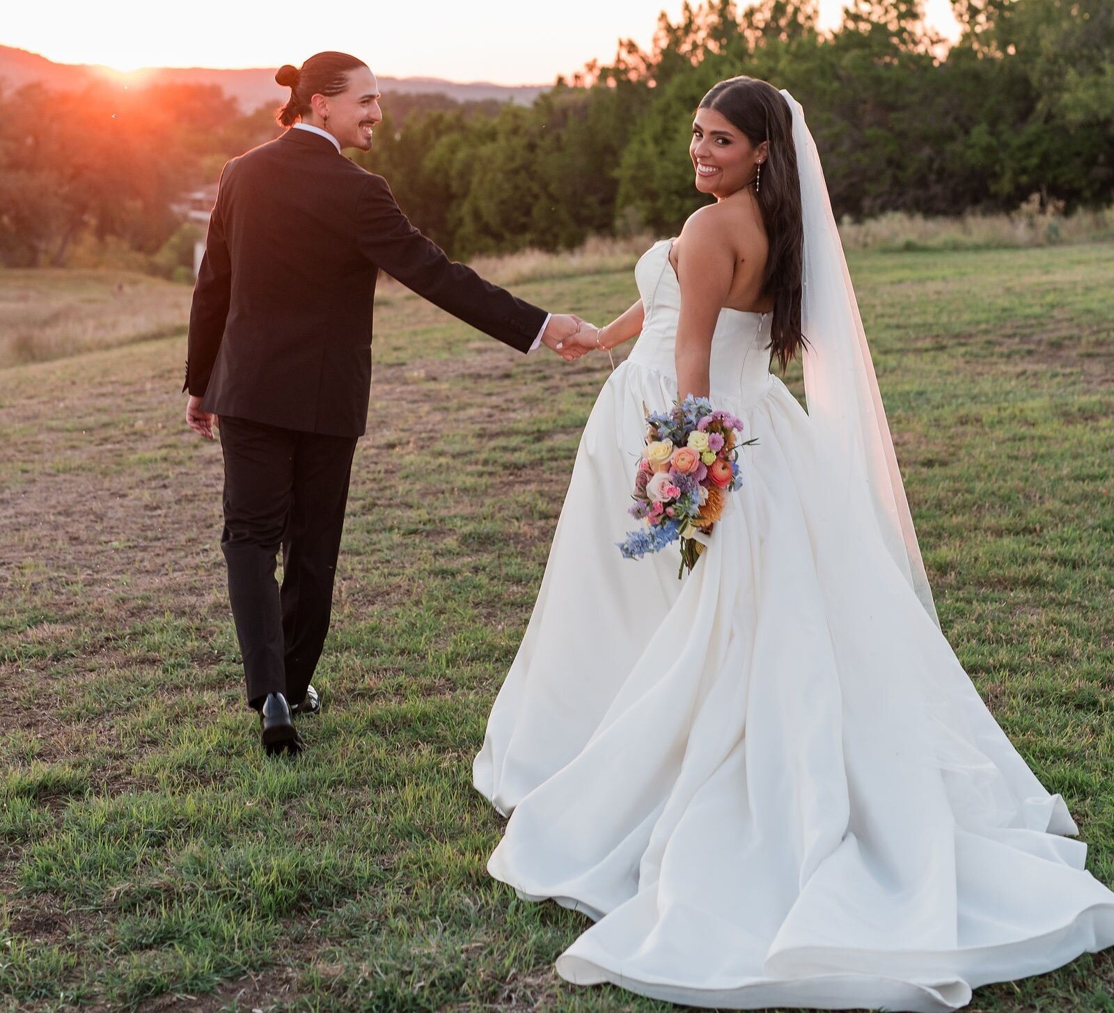 The smiling bride and groom walk hand-in-hand through a grassy field during golden hour sunset.