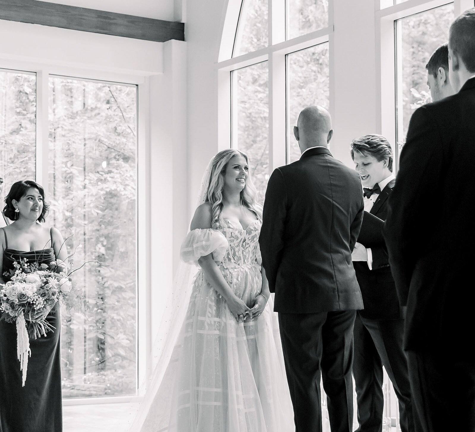 A black and white photo of a bride smiling at the groom during an indoor wedding ceremony with tall windows.