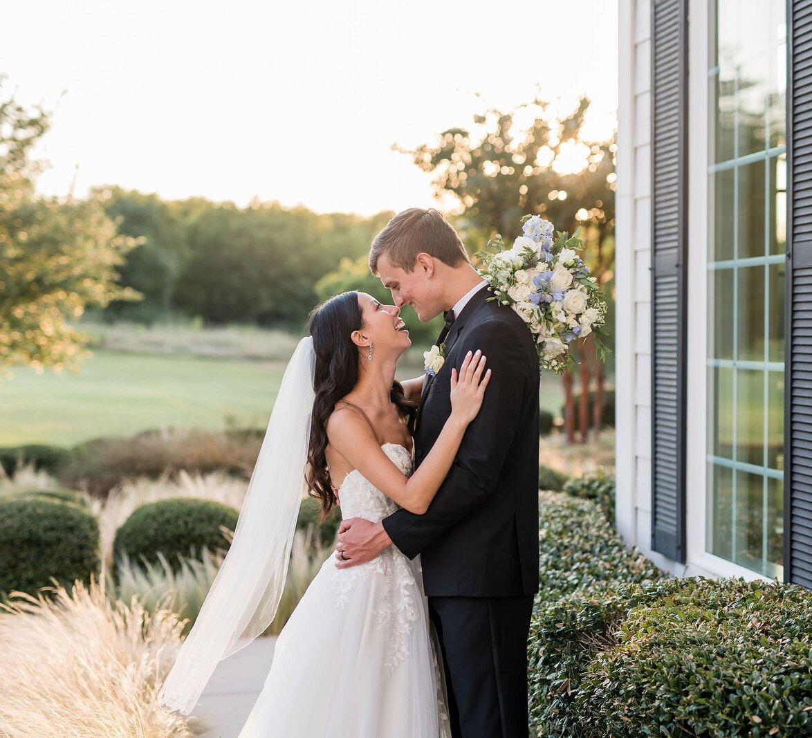 A candid portrait of the newlywed bride and groom embracing and smiling next to a white house at sunset.