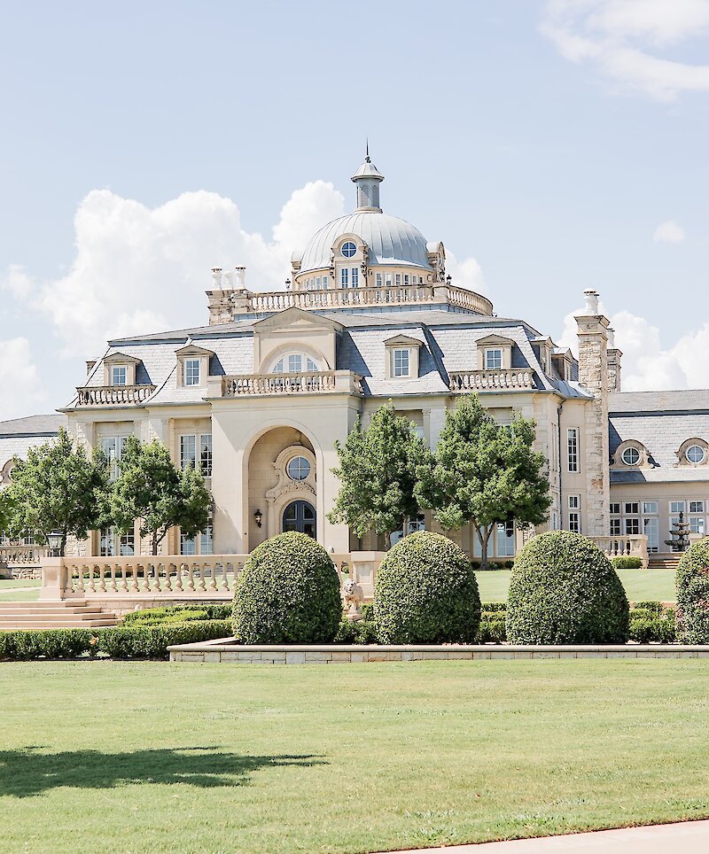 The stunning exterior of a luxury wedding and event venue features a light stone facade, a domed roof, and manicured green lawns and hedges.
