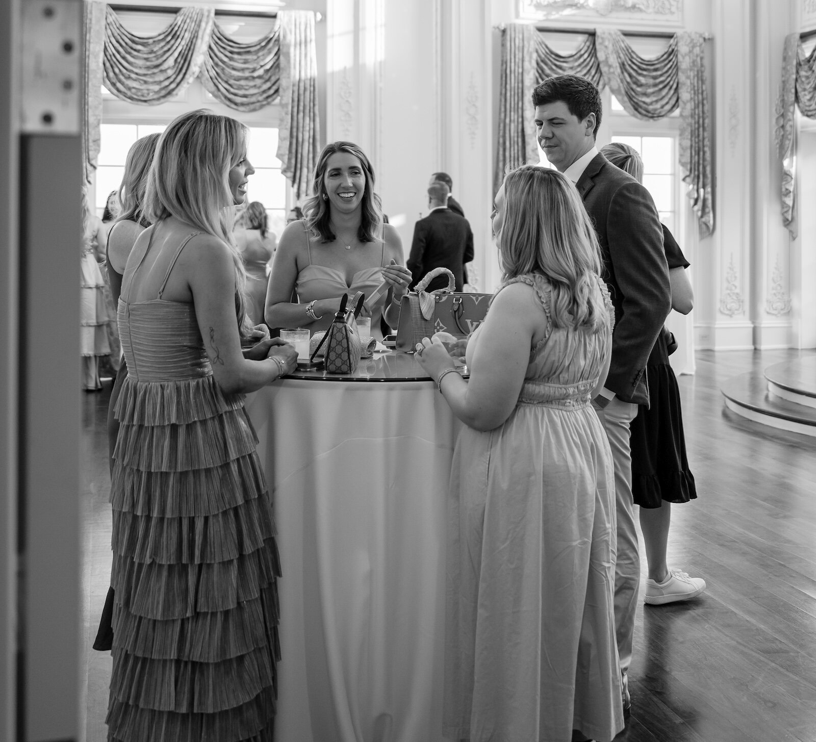 A black and white photo of guests at a celebration socializing around a cocktail table in a grand ballroom with large windows and a crystal chandelier.