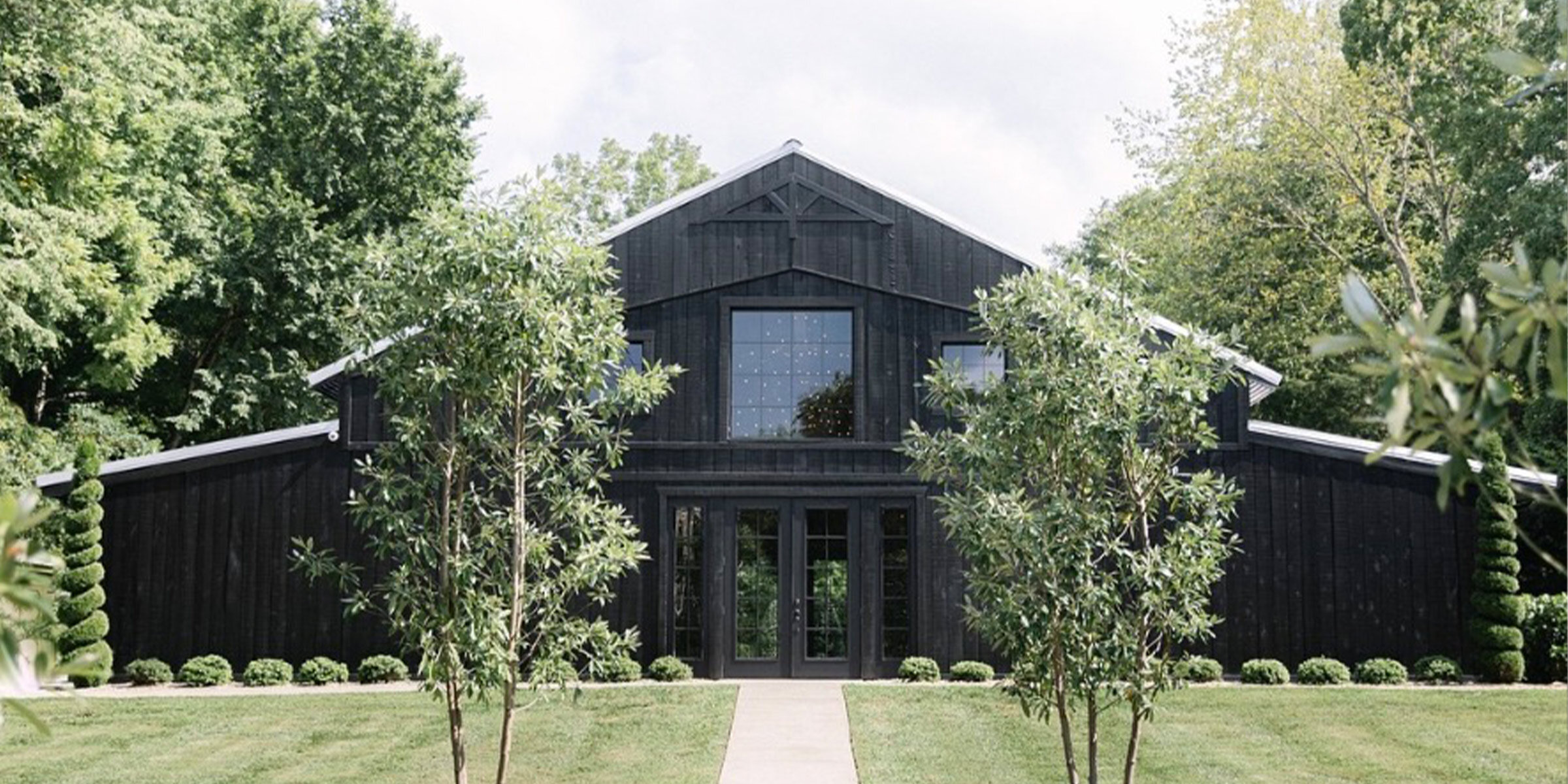 Exterior of a sprawling rustic wedding venue with a low-slung shingled roof, light stone facade, exposed wooden beams on the covered entryway, and native landscaping in the foreground.