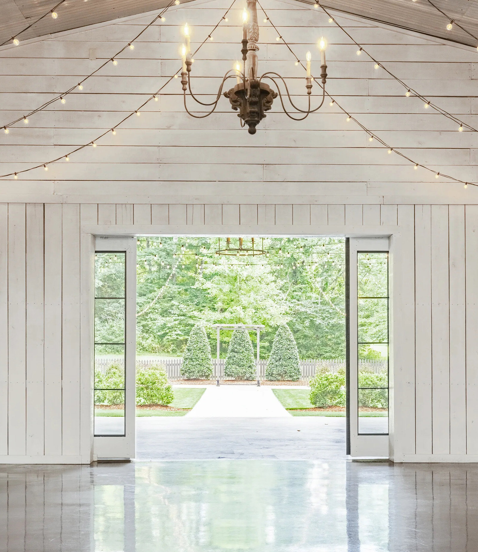 white barn reception hall looking out to green nature with string lights and chandelier