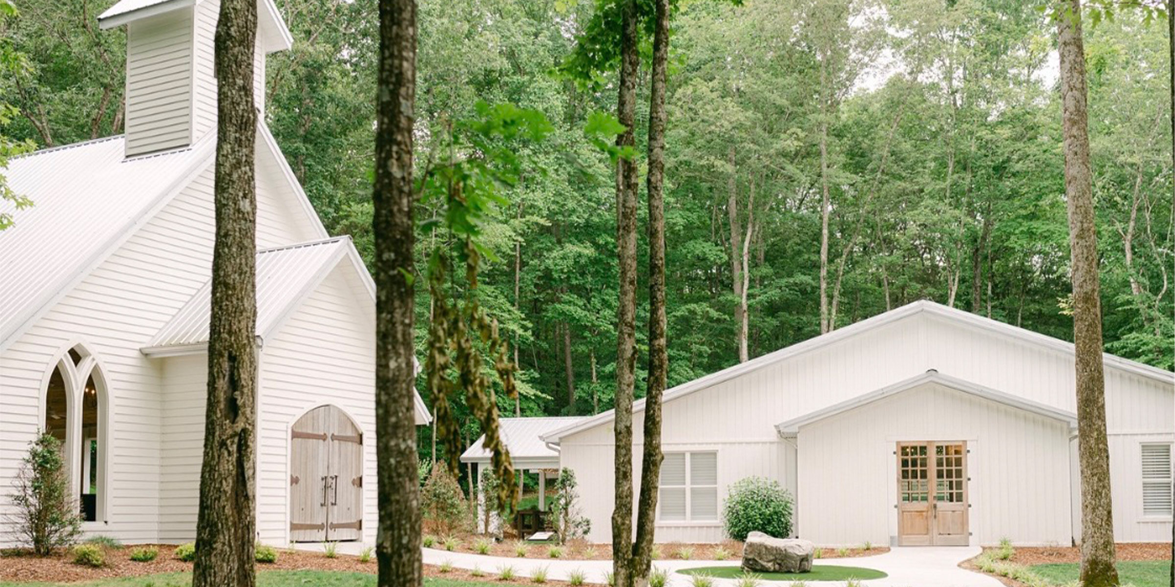 Exterior of a sprawling rustic wedding venue with a low-slung shingled roof, light stone facade, exposed wooden beams on the covered entryway, and native landscaping in the foreground.