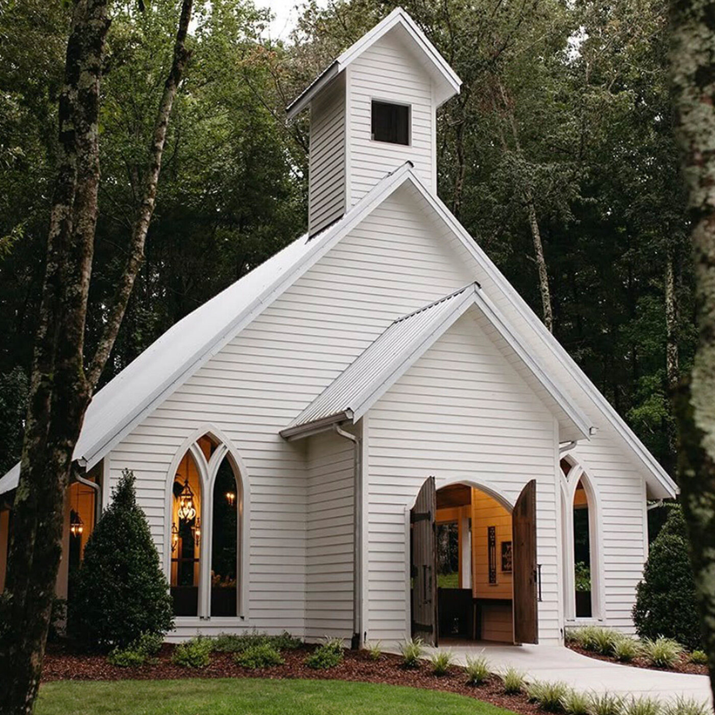 Outdoor wedding ceremony setup featuring a wooden pavilion altar with exposed beams, rows of white folding chairs, a flagstone aisle, and floral arrangements of white and blue hydrangeas, all set beneath large oak trees.