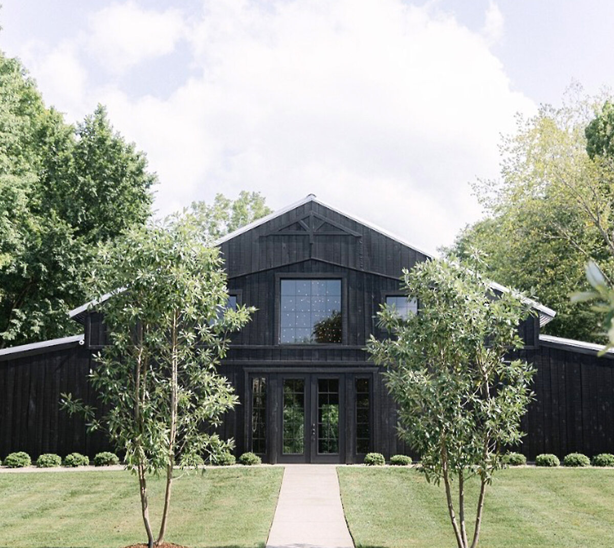 A rendered view of the modern rustic wedding venue exterior at dusk, featuring gray wood siding, a stone entrance, and a covered drive-up area with cars and wild landscaping.