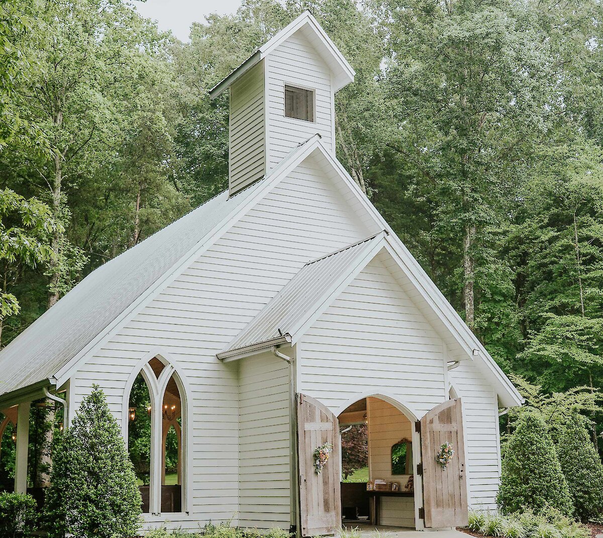 A rendered view of the modern rustic wedding venue exterior at dusk, featuring gray wood siding, a stone entrance, and a covered drive-up area with cars and wild landscaping.