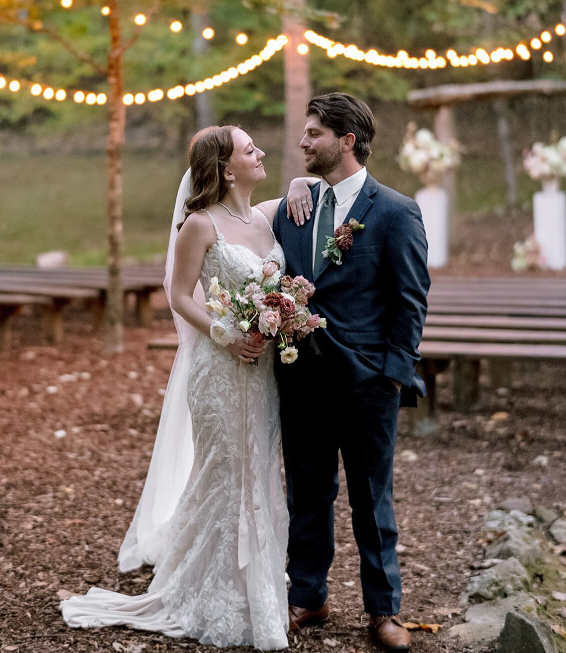 bride and groom standing under outdoor ceremony arch with tall trees and string lights