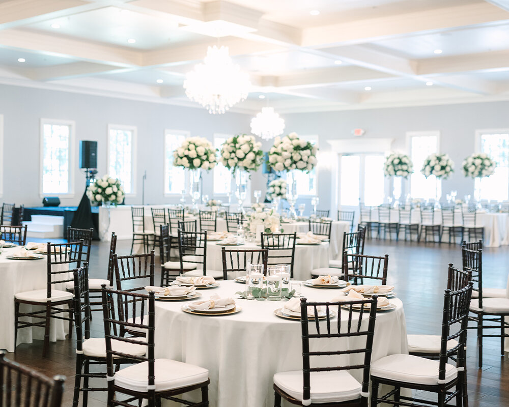 A classic wedding reception ballroom set up with white-linen covered round tables and dark Chiavari chairs.