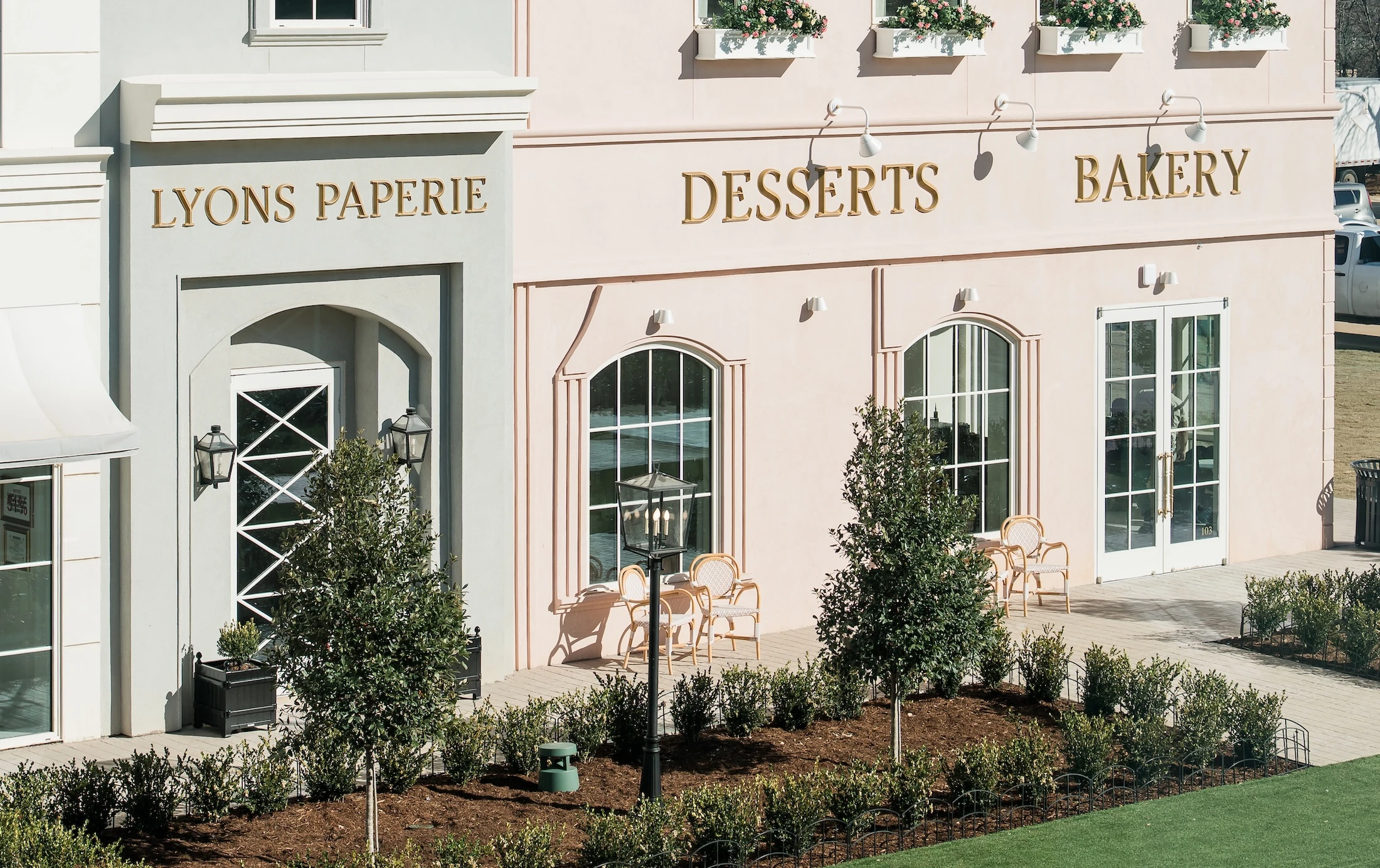 The picturesque exterior of a European-style venue with a light pink facade, striped awnings, and gold lettering for "Desserts Bakery" and "Lyons Paperie."