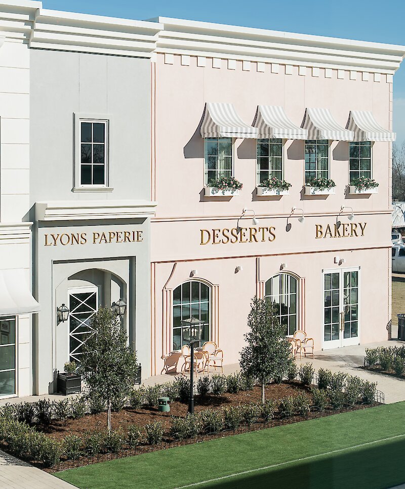 The picturesque exterior of a European-style venue with a light pink facade, striped awnings, and gold lettering for "Desserts Bakery" and "Lyons Paperie."