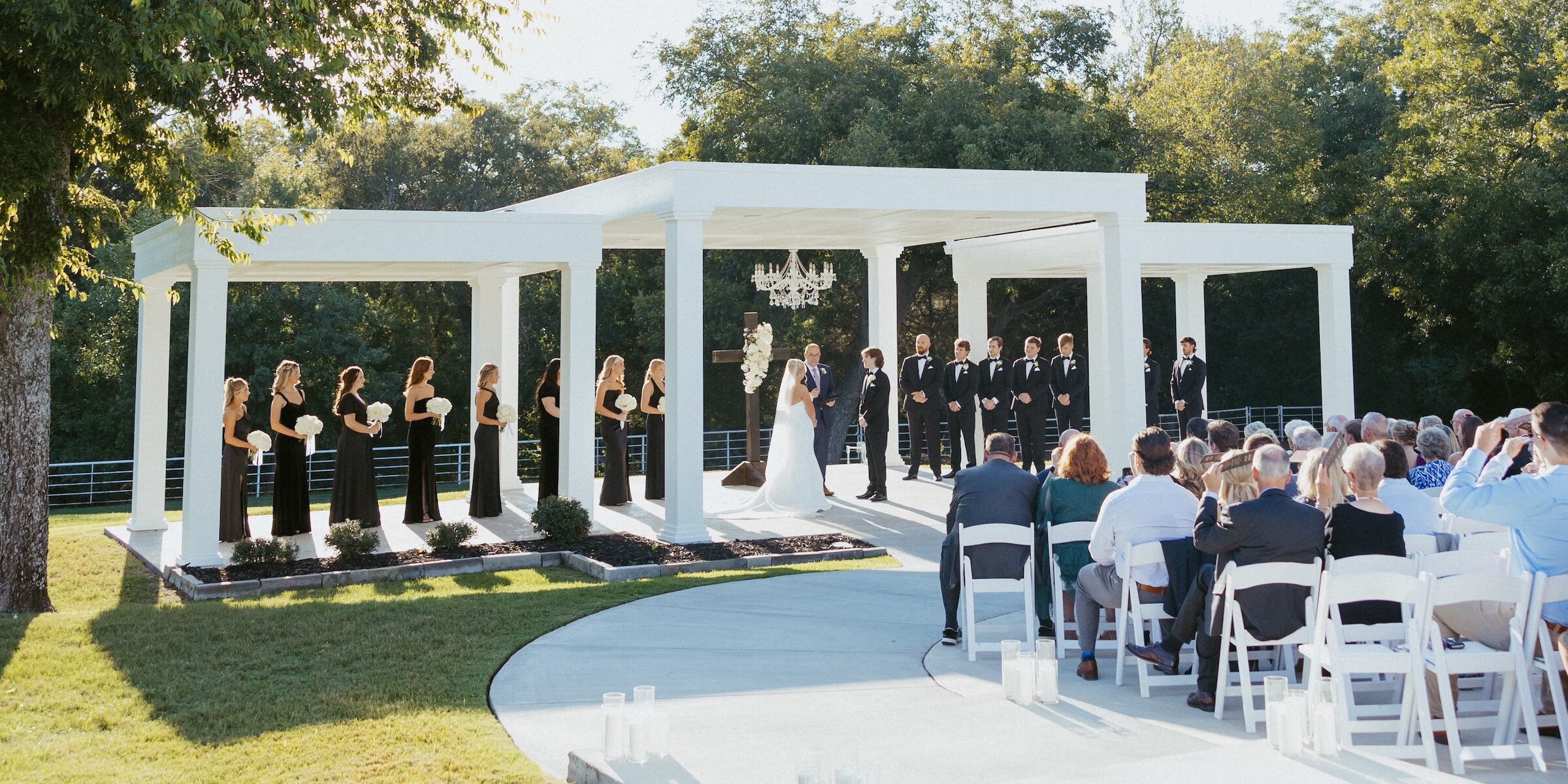 An outdoor wedding ceremony under a white columned pergola with a crystal chandelier, featuring the bridal party in black attire and seated guests.