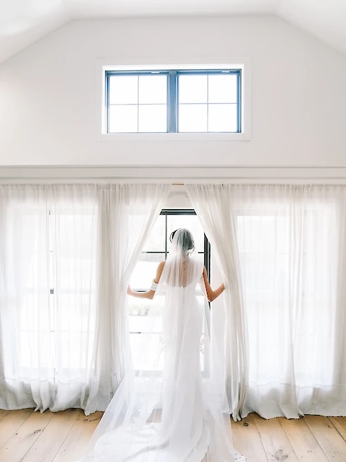 The bride stands in a white gown and long veil, looking out of a large window, as she parts sheer white drapes in a bright, modern room.