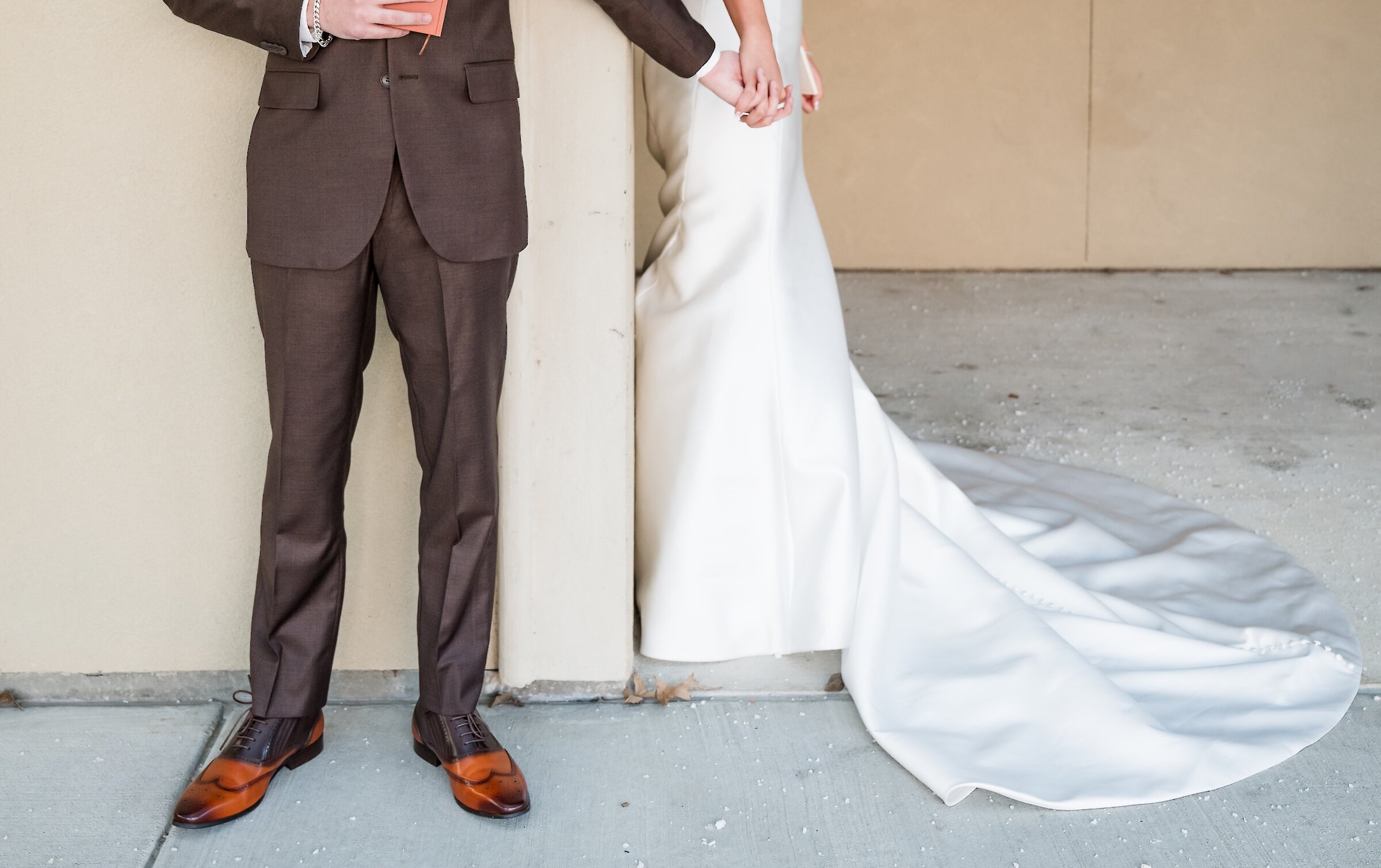 The smiling groom in a brown suit holds the hand of his bride, who stands around a corner, for a first touch before their ceremony.