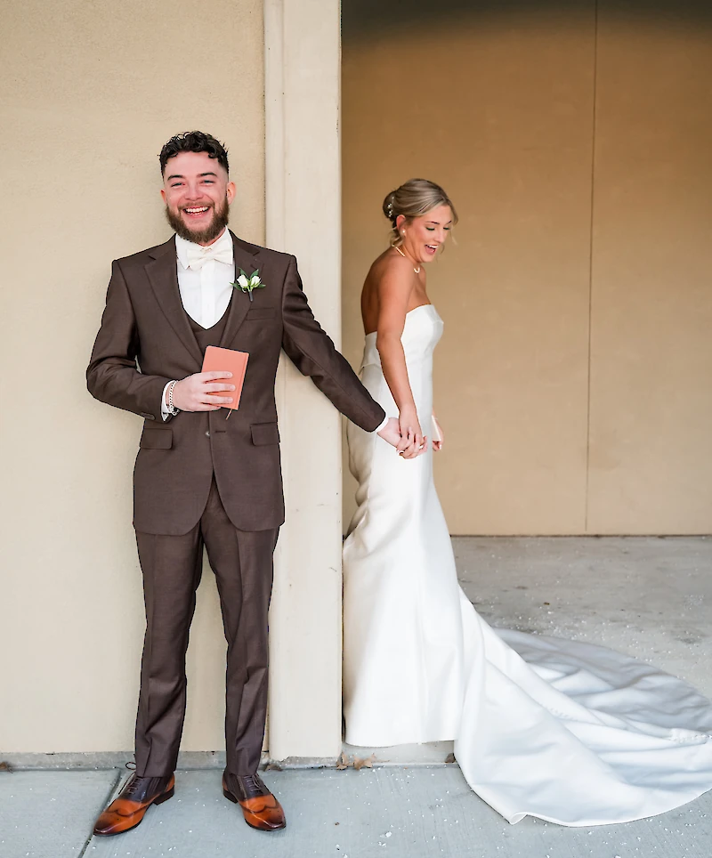 The smiling groom in a brown suit holds the hand of his bride, who stands around a corner, for a first touch before their ceremony.
