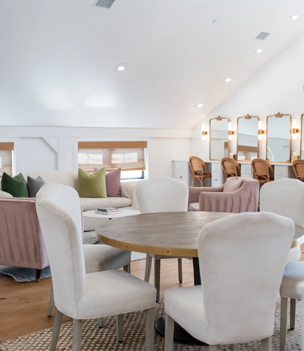 A bright bridal suite featuring a comfortable lounge area and a professional makeup vanity station with four ornate gold mirrors and wicker chairs.