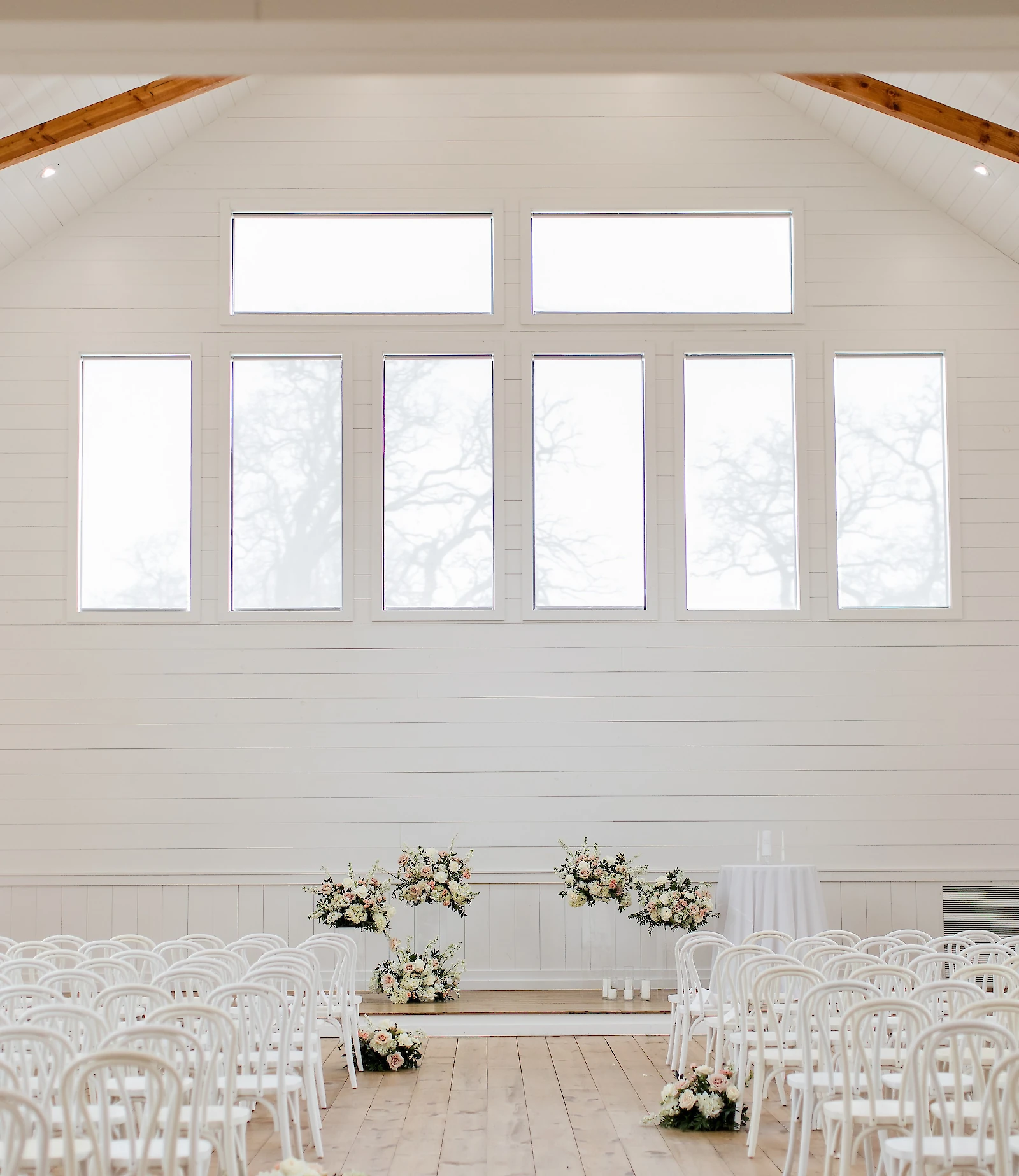 A bright, empty white barn chapel interior with vaulted ceiling, large rectangular windows, and white chairs set for a ceremony with white and blush floral arrangements.