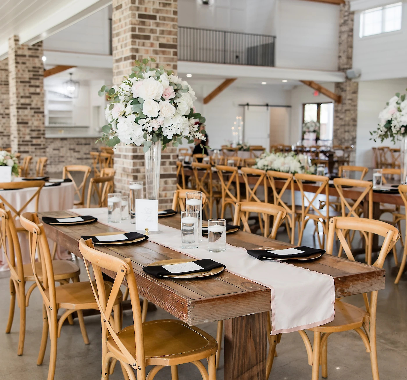 A long, rustic wooden farmhouse table is set with blush runners, black napkins, and a tall white and pink floral centerpiece in a barn reception venue.