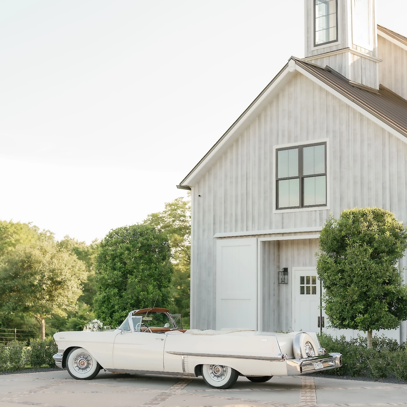 A creamy white vintage convertible car is parked next to the white, modern farmhouse barn exit under a bright sky.