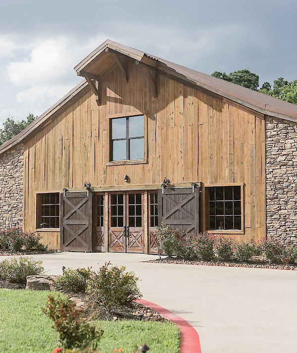 Rustic wooden barn-style venue with stone accents, large sliding barn doors, and blooming landscaping along a curved driveway under a cloudy sky.