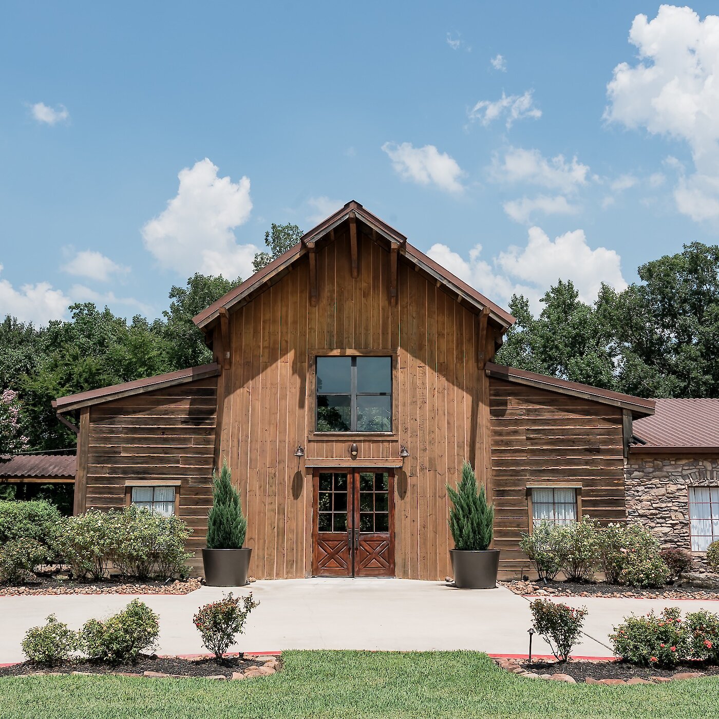 Exterior of a rustic wood and stone barn wedding venue with a central gable entrance and wooden double doors, surrounded by lush green trees and landscaping under a blue, cloudy sky.