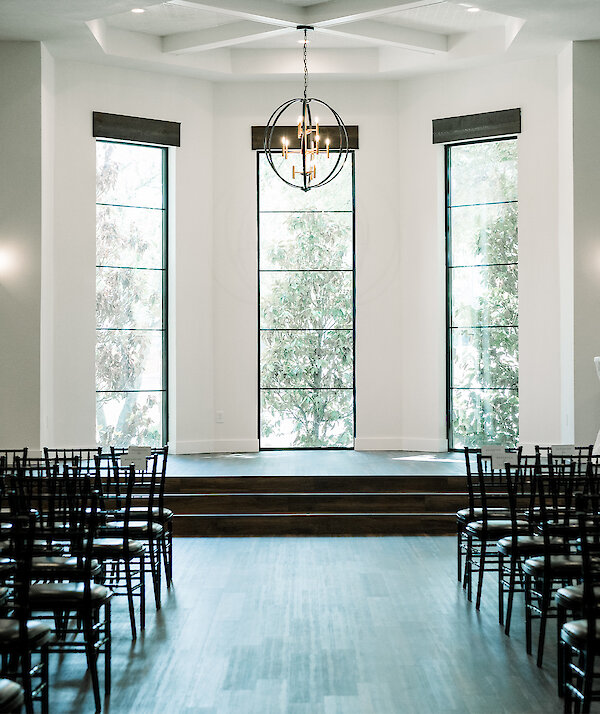 Covered stone entrance with lantern-style wall lights, black double doors, and tall trees creating dappled shade around the walkway.