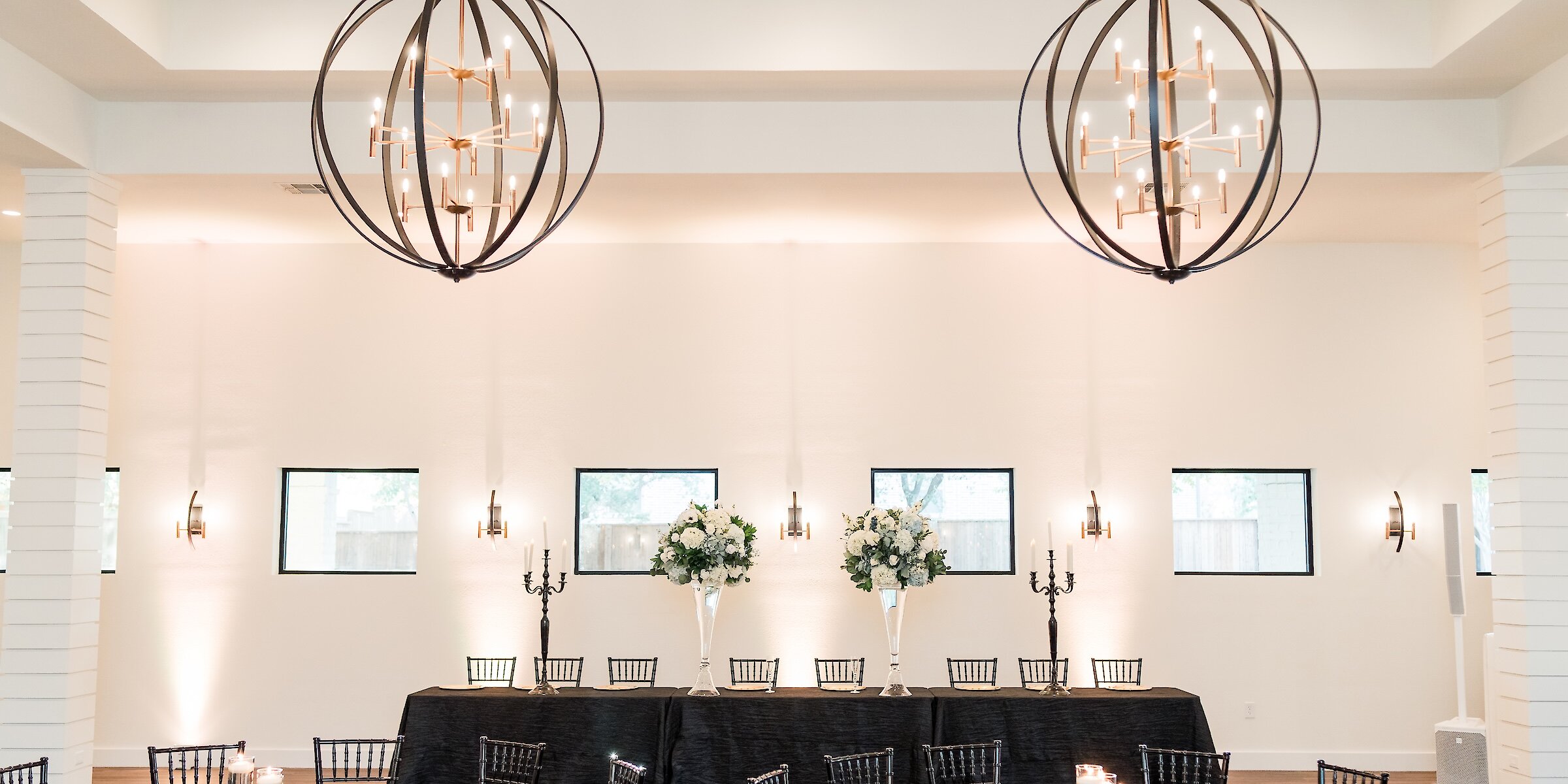 Modern wedding reception hall featuring a dramatic black and gold color scheme. Round tables and a long head table are set with black linens, black Chiavari chairs, gold charger plates, and tall white floral centerpieces beneath large, contemporary orb chandeliers.