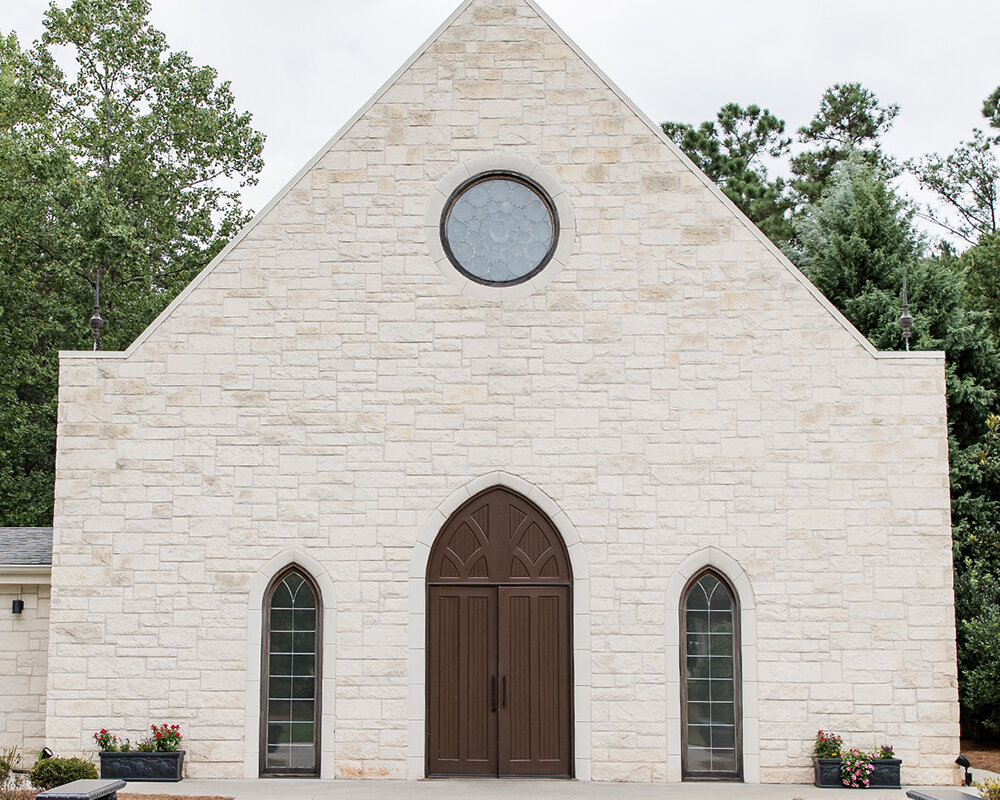 The gabled exterior of a wedding chapel made of light-colored limestone blocks with a round stained-glass window above a dark wood double-door entrance.