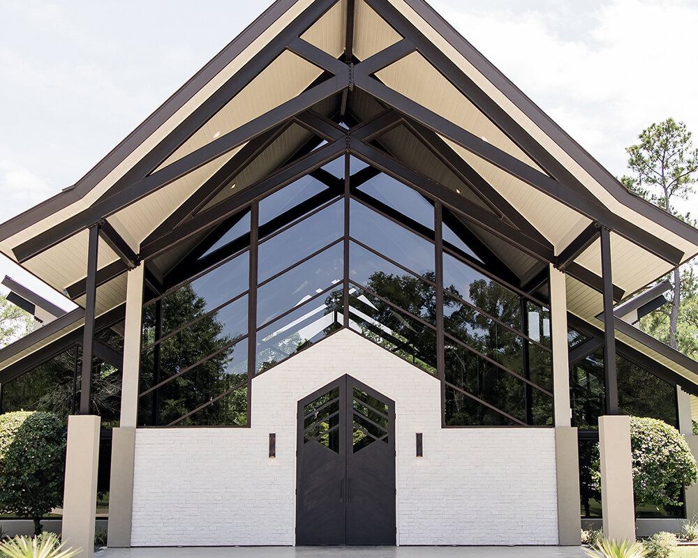 The striking modern exterior of an event venue with large black-framed windows, a slanted roofline, and a welcome sign near the entrance.