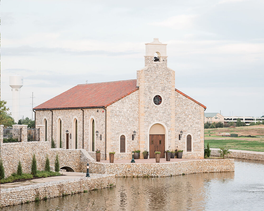 A beautiful stone chapel with a bell tower and red tile roof sits on a stone retaining wall overlooking a pond.