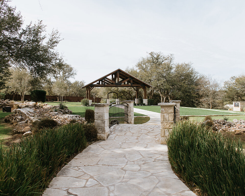 An outdoor wedding venue with a stone path leading over a bridge to a wooden covered pavilion on a grassy lawn.
