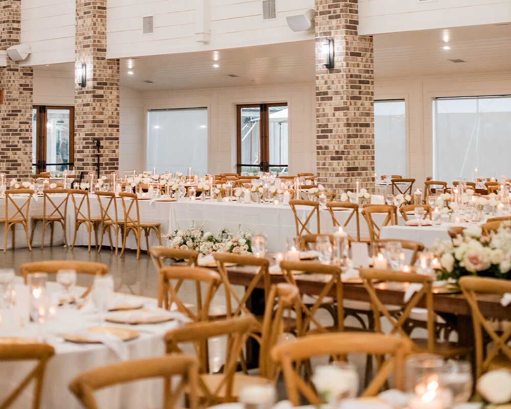 An elegant wedding reception dinner setup with cross-back chairs, white linens, and tables decorated with candles and white floral arrangements.