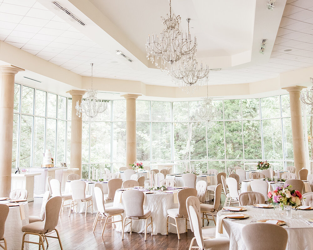 An elegant wedding reception hall featuring a circular wall of windows, crystal chandeliers, large round tables with white linens, and light beige chairs on a wood floor.