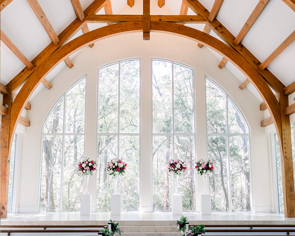 The interior of a rustic modern wedding chapel with wooden arched beams and large windows overlooking trees.