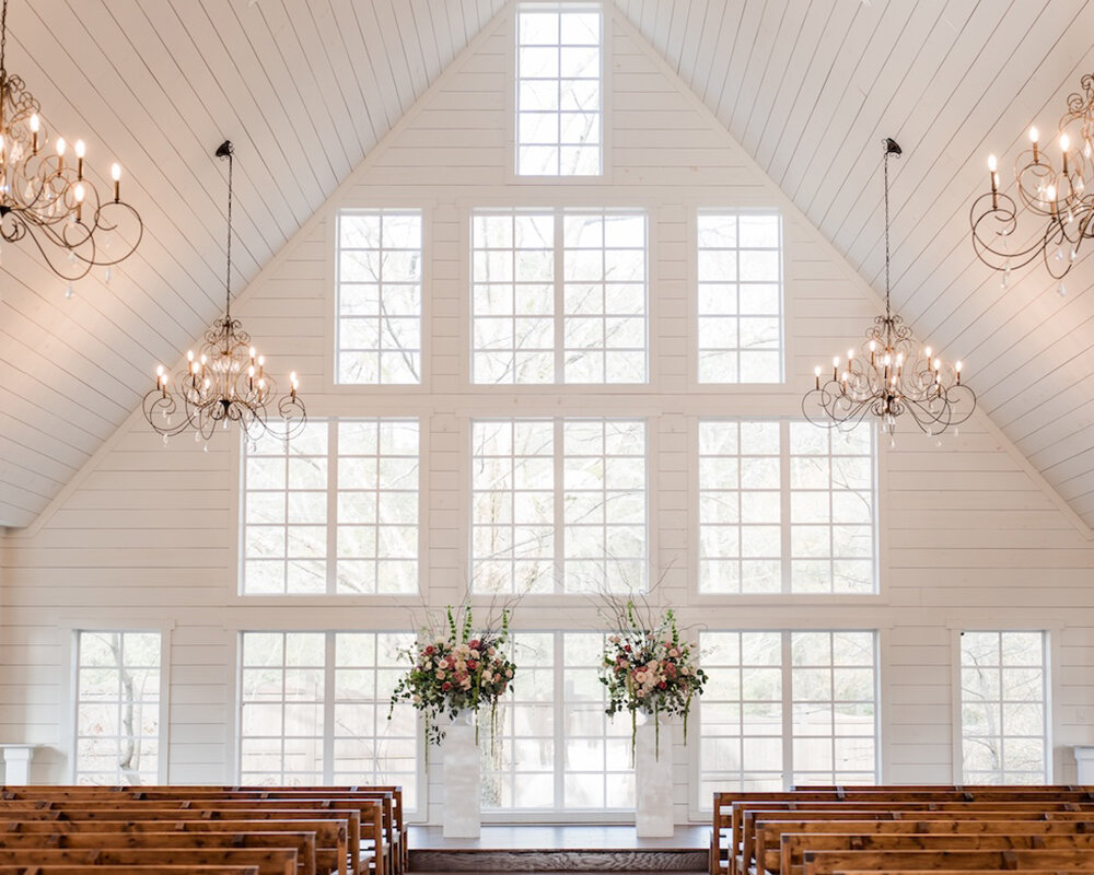 The bright, airy interior of a white chapel with high ceilings, many windows, elegant chandeliers, and dark wooden pews.