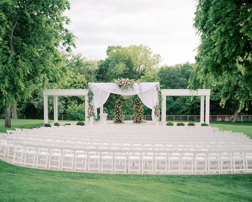 A beautiful outdoor wedding ceremony setup with white folding chairs arranged in a semi-circle facing a white draped arbor with floral accents.